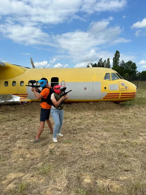 Deux joueurs de paintball masqués en action devant l'avion, sur le terrain de Happy Forest